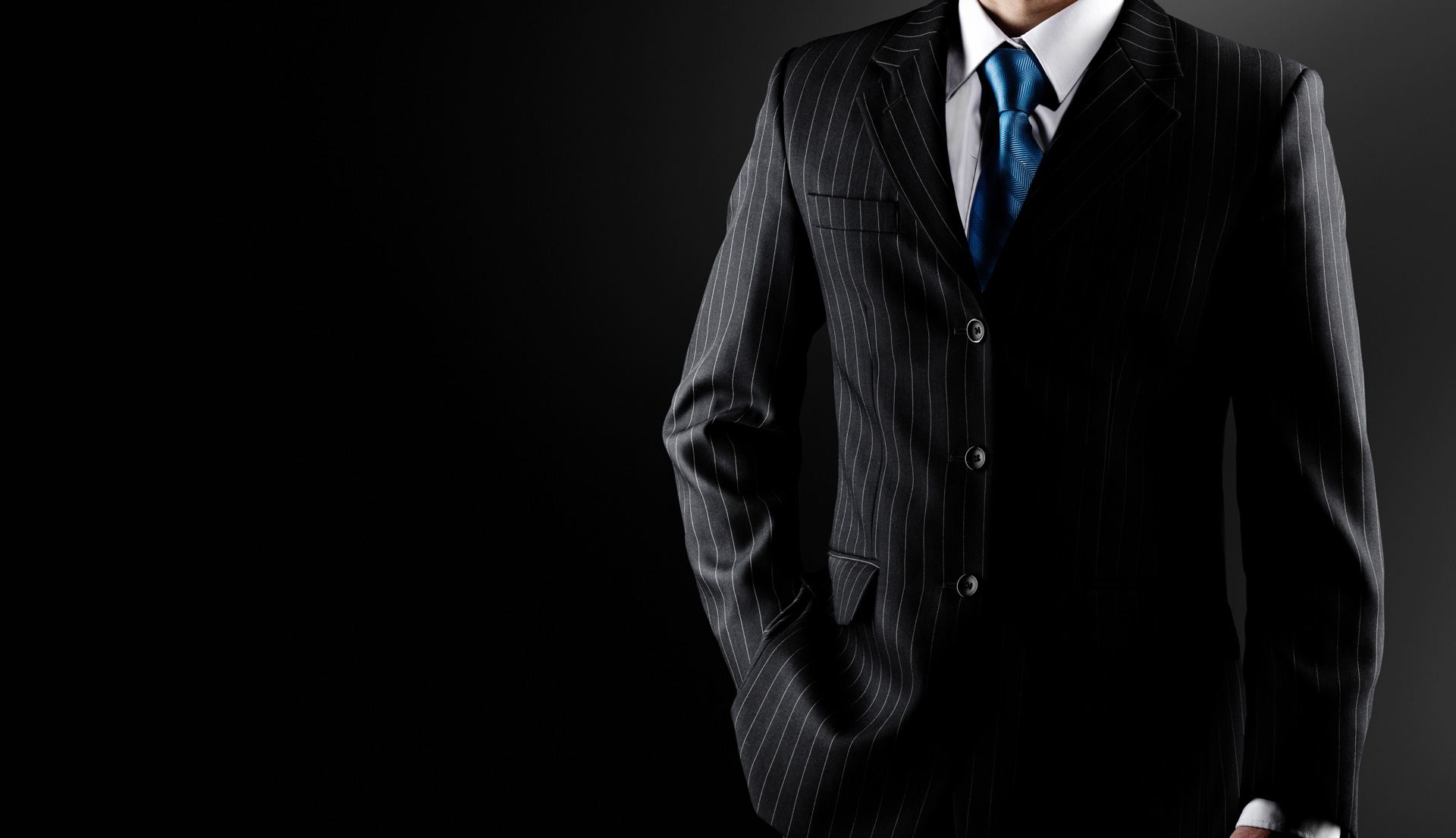 A man in a dark pinstripe suit and blue tie, photographed low-key against black